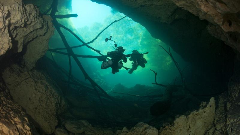 Two divers descend down into an underwater cave, backlit by a blue-green light